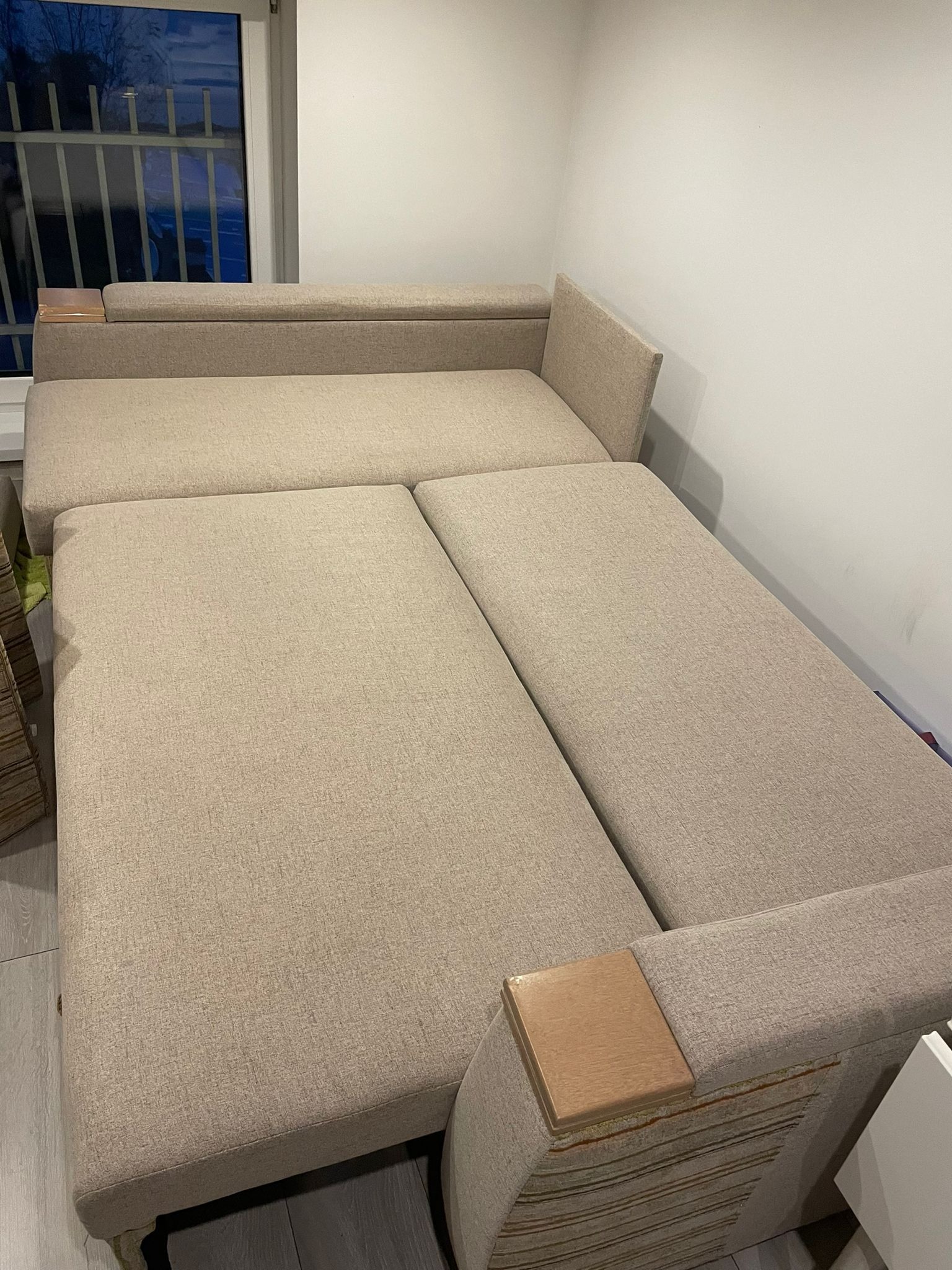 Beżowa sofa modułowa z drewnianymi akcentami, rozłożona, widoczna w pomieszczeniu z oknem i białą ścianą.