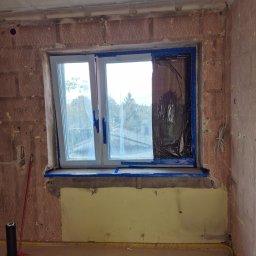 ARKADIUSZ Grzegorz Płatek - Pokój w trakcie przygotowań do malowania: okno zabezpieczone folią i taśmą malarską, ściany z resztkami tapety, żółta farba na dolnej części ściany, widoczna skrzynka narzędziowa i rolka folii.