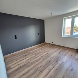 ARKADIUSZ Grzegorz Płatek - Pusty pokój po remoncie z drewnopodobną podłogą, szarą ścianą akcentującą i białymi ścianami bocznymi, widoczne gniazdka elektryczne i okno z widokiem na drzewa.