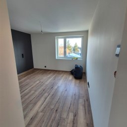 ARKADIUSZ Grzegorz Płatek - Wnętrze pokoju po remoncie z ułożonymi panelami podłogowymi, pomalowaną jedną ścianą na ciemny kolor, workiem z odpadami w rogu i widocznym przez okno fragmentem osiedla.