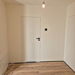 ARKADIUSZ Grzegorz Płatek - Wnętrze po remoncie w Myszkowie: świeżo ułożone panele podłogowe w odcieniach brązu, białe ściany i minimalistyczne białe drzwi z czarnymi detalami, surowa żarówka zwisająca z sufitu.