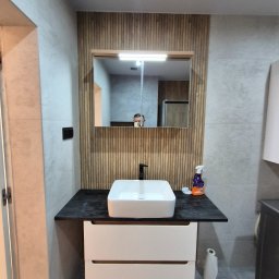 ARKADIUSZ Grzegorz Płatek - Nowoczesna łazienka z kwadratową umywalką nablatową, czarnym blatem, drewnianymi panelami za lustrem i szarą płytką na ścianach oraz podłodze. W lustrze odbija się osoba robiąca zdjęcie.