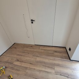 ARKADIUSZ Grzegorz Płatek - Ułożone panele podłogowe w pokoju z białymi ścianami i czarnymi listwami przypodłogowymi, widoczne narzędzia montażowe w rogu.