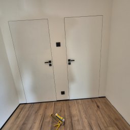 ARKADIUSZ Grzegorz Płatek - Dwie białe, minimalistyczne drzwi wewnętrzne z czarnymi klamkami i ukrytymi ościeżnicami, zamontowane w skosie poddasza, na podłodze z paneli leży pistolet do silikonu i tuba z silikonem.