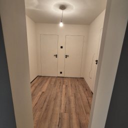ARKADIUSZ Grzegorz Płatek - Korytarz po remoncie z drewnopodobną podłogą, białymi ścianami i dwiema białymi, minimalistycznymi drzwiami z czarnymi klamkami, oświetlony pojedynczą żarówką