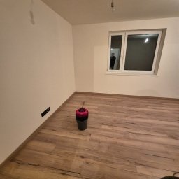ARKADIUSZ Grzegorz Płatek - Pokój po położeniu paneli podłogowych z widocznym wiadrem i mopem, surową żarówką oraz odbiciem osoby w oknie.