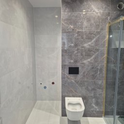 ARKADIUSZ Grzegorz Płatek - Nowoczesna łazienka z szarymi płytkami imitującymi marmur, podwieszana toaleta z czarnym przyciskiem spłukiwania, rolka papieru toaletowego na podłodze, kabina prysznicowa z elementami w kolorze...
