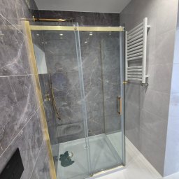 ARKADIUSZ Grzegorz Płatek - Nowoczesna kabina prysznicowa z przesuwnymi drzwiami, złotymi ramami i deszczownicą, na tle szarych płytek imitujących marmur, z białym grzejnikiem drabinkowym po prawej stronie.