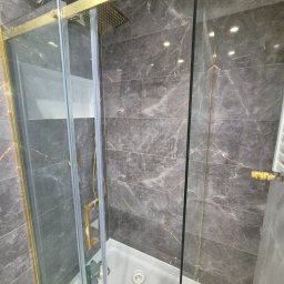 ARKADIUSZ Grzegorz Płatek - Szklana kabina prysznicowa z brodzikiem, złotymi ramami i deszczownicą, na tle marmurowych płytek w odcieniach szarości z delikatnym złotym żyłowaniem. Widoczny odpływ liniowy i fragment grzejnika.