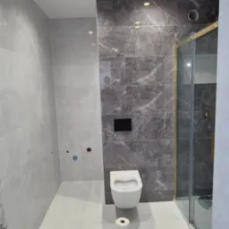 Nowoczesna łazienka z szarymi płytkami imitującymi marmur, podwieszana toaleta i kabina prysznicowa ze złotymi profilami; widoczny papier toaletowy na podłodze.