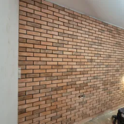 Ściana z cegieł dekoracyjnych w odcieniach brązu i beżu, z widocznymi puszkami elektrycznymi i prowizorycznym oświetleniem, prawdopodobnie w trakcie prac wykończeniowych w Myszkowie. W rogu...