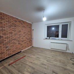 ARKADIUSZ Grzegorz Płatek - Pokój w trakcie prac wykończeniowych: ściana z cegły klinkierowej, panele podłogowe w odcieniach brązu, grzejnik pod oknem, wisząca żarówka i poziomica na podłodze.