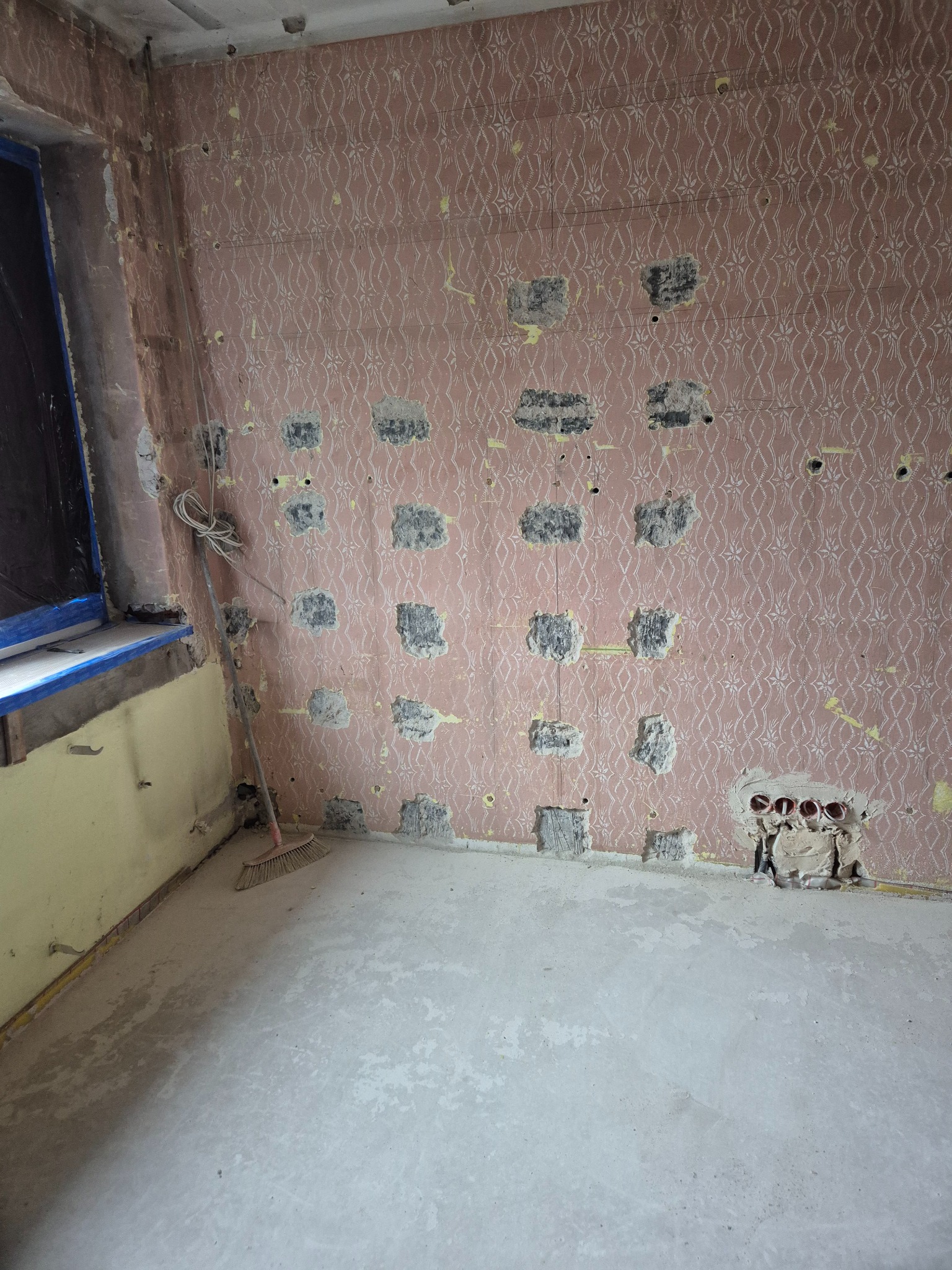 Pokój w trakcie przygotowań do prac wykończeniowych: ściana z resztkami różowej tapety w kwiaty, liczne otwory po usuniętych gniazdkach i fragmentach tynku, widoczne przewody elektryczne, okno...