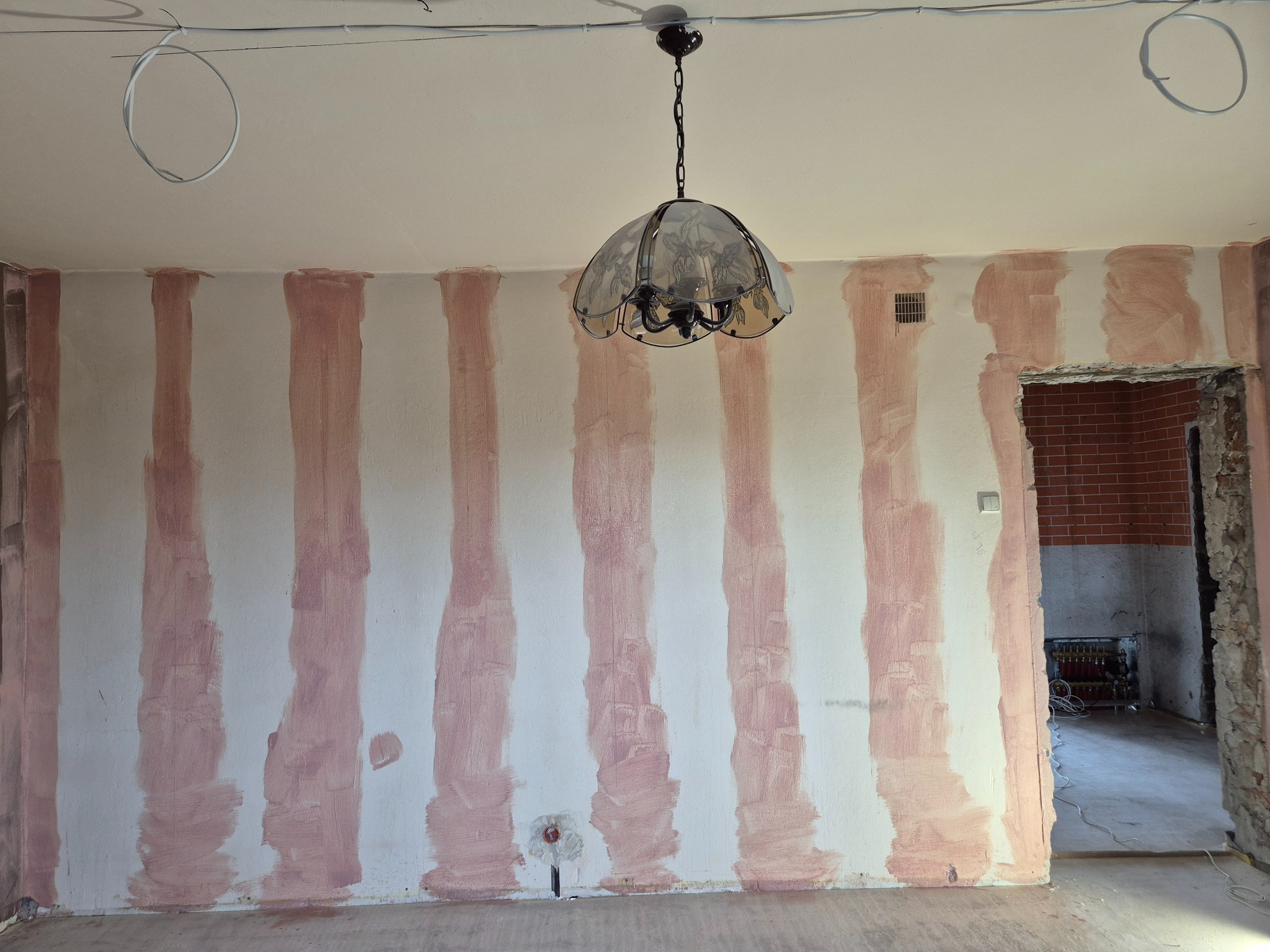 Pokój w trakcie przygotowania do malowania: ściany z pionowymi pasami zagruntowane, widoczna instalacja elektryczna na suficie, wisząca lampa i fragment sąsiedniego pomieszczenia z instalacją...