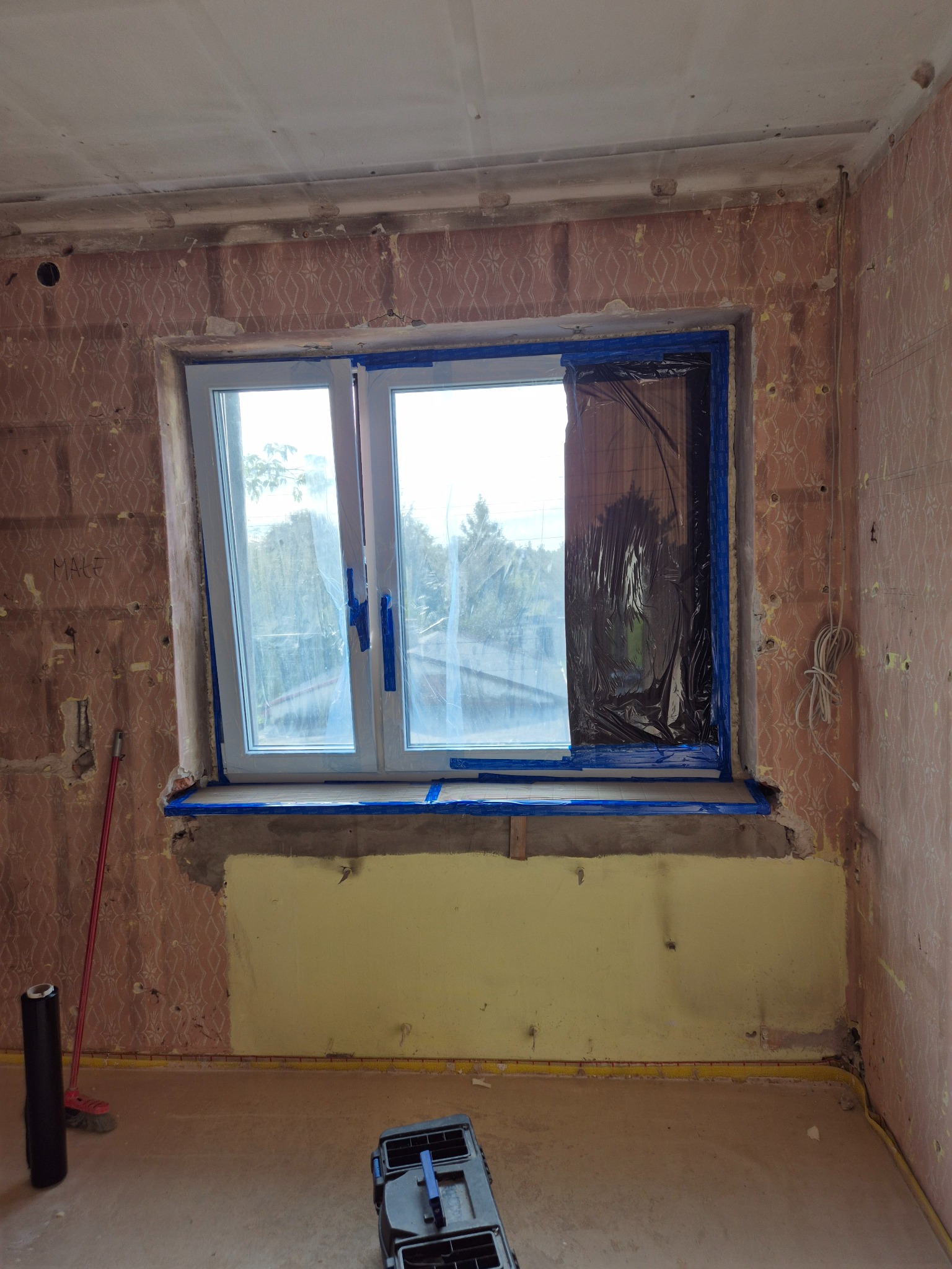 Pokój w trakcie przygotowań do malowania: okno zabezpieczone folią i taśmą malarską, ściany z resztkami tapety, żółta farba na dolnej części ściany, widoczna skrzynka narzędziowa i rolka folii.