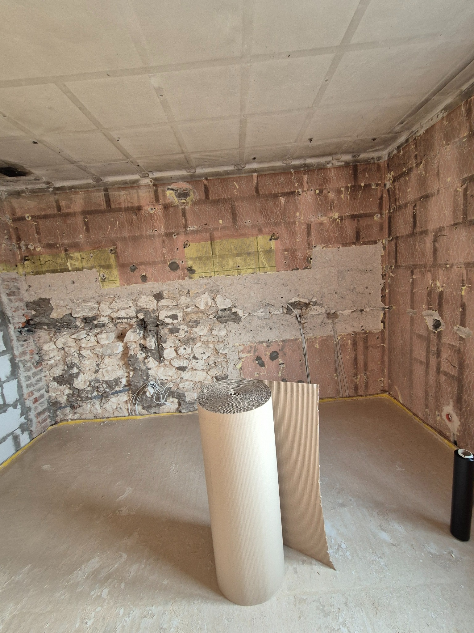Wnętrze w trakcie prac przygotowawczych: odsłonięte mury, częściowo usunięta tapeta, widoczne przewody elektryczne, rolka i arkusz tektury falistej do zabezpieczenia podłogi, czarna tuba na podłodze.