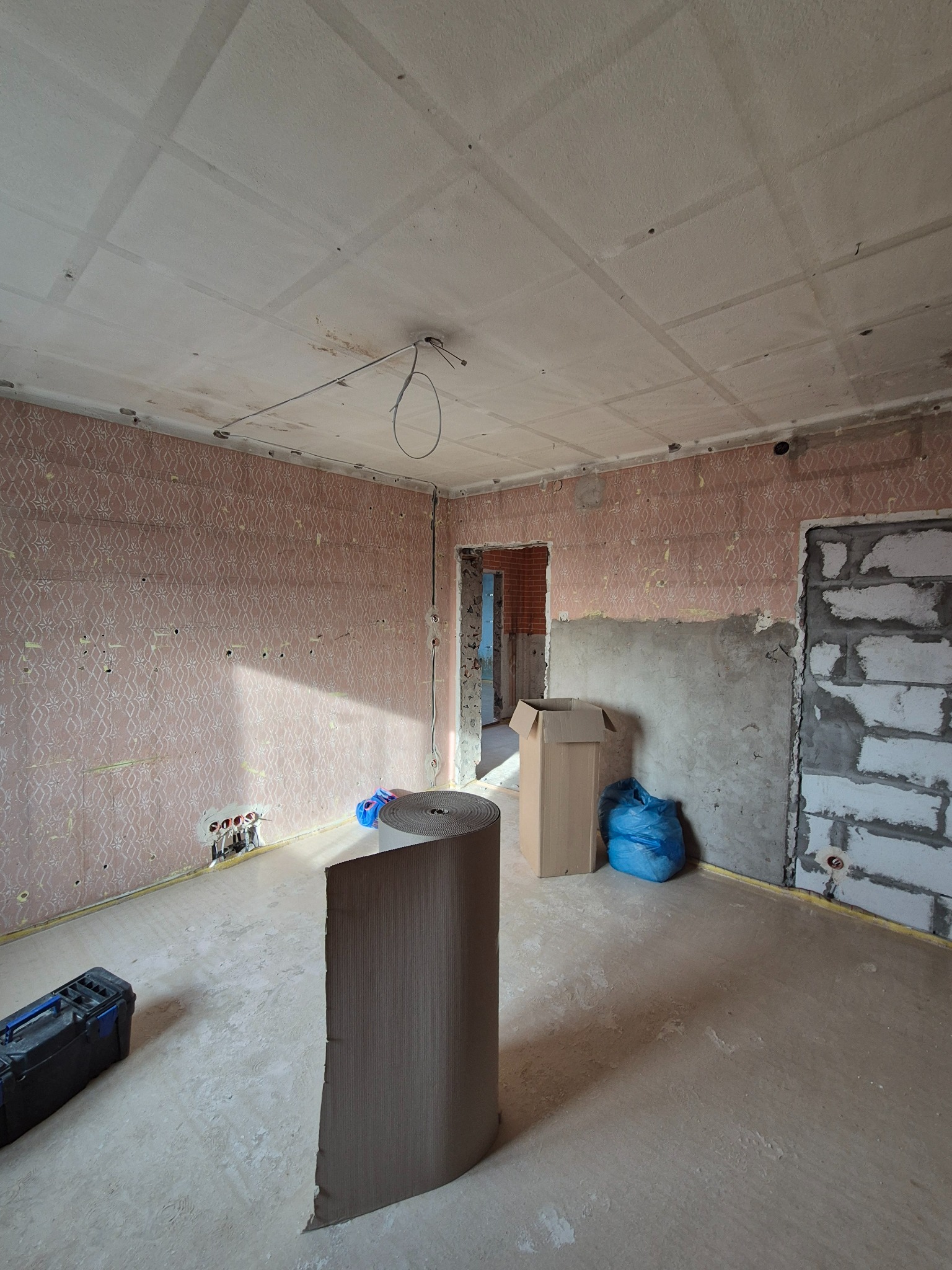 Pokój w trakcie prac wykończeniowych: ściany z fragmentami starej tapety i surowego tynku, widoczne instalacje elektryczne, zwój materiału podłogowego, skrzynka z narzędziami, oraz fragment muru...