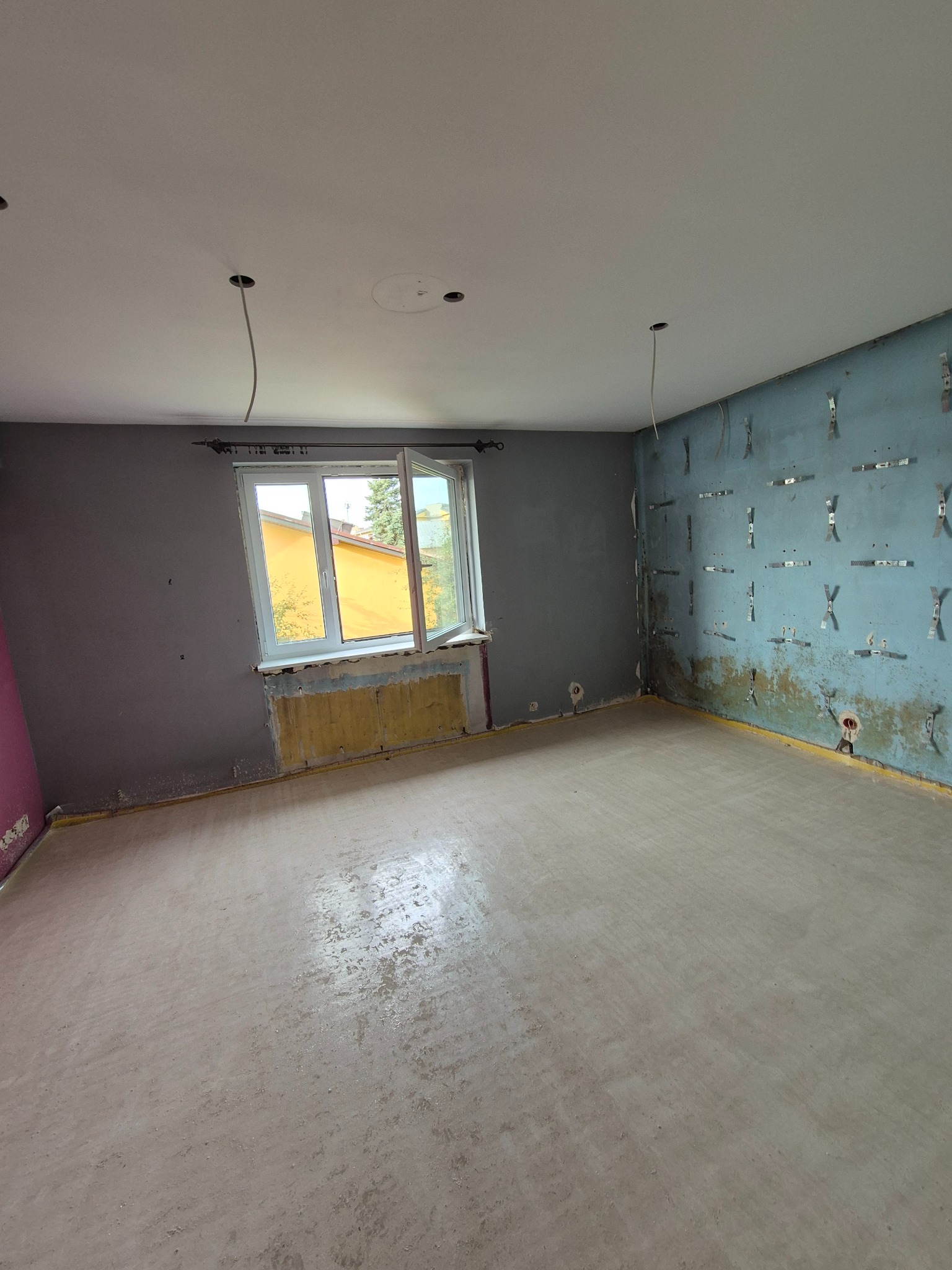 Pokój w trakcie przygotowań do prac wykończeniowych: ściany częściowo przygotowane pod montaż płyt kartonowo-gipsowych z widocznymi profilami, okno otwarte, odsłonięty grzejnik, podłoga z wylewką...