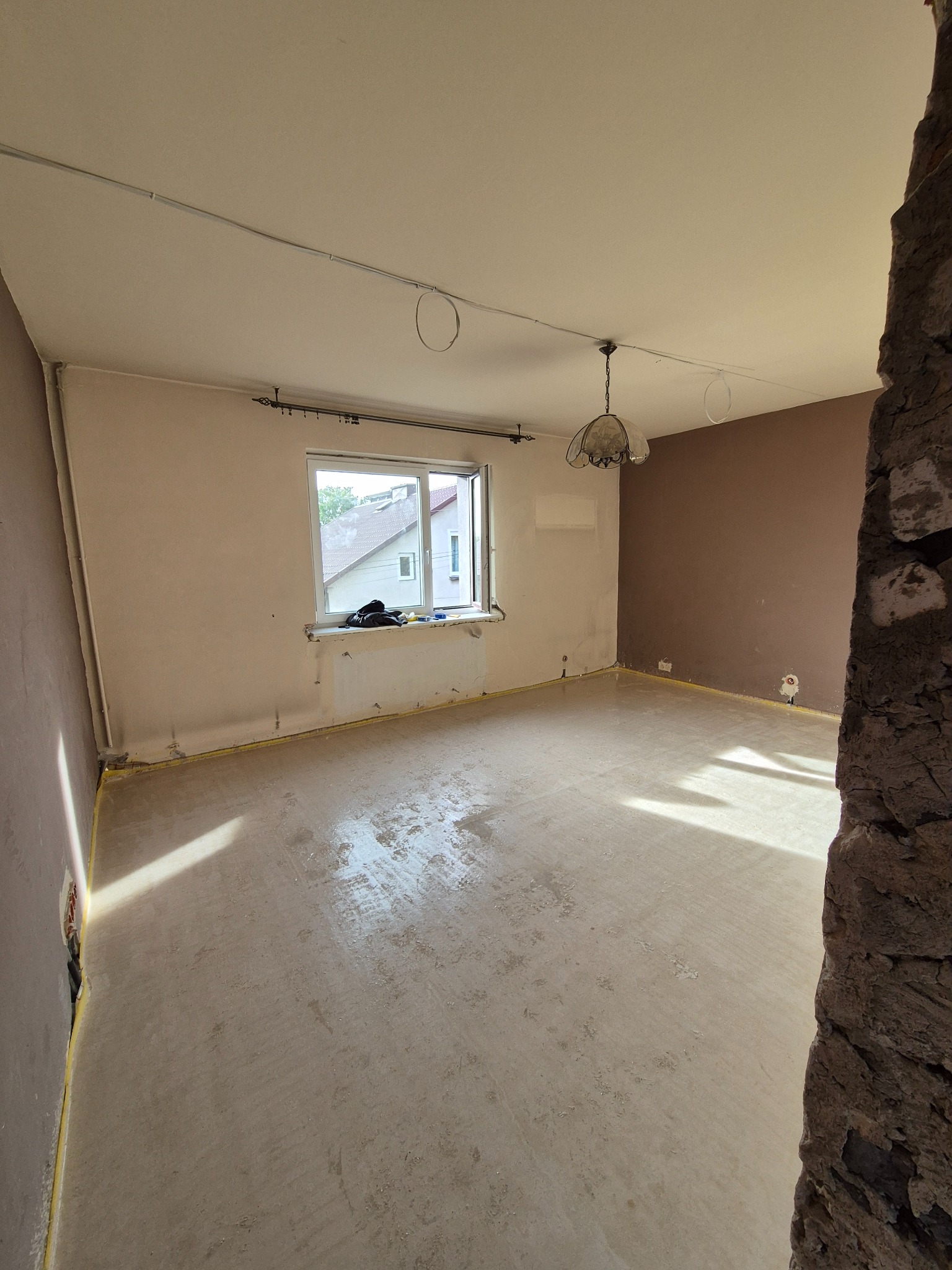 Pokój w trakcie remontu: przygotowana podłoga, częściowo pomalowane ściany w odcieniach beżu i brązu, widoczne instalacje elektryczne na suficie i zabezpieczenia folią malarską przy podłodze,...