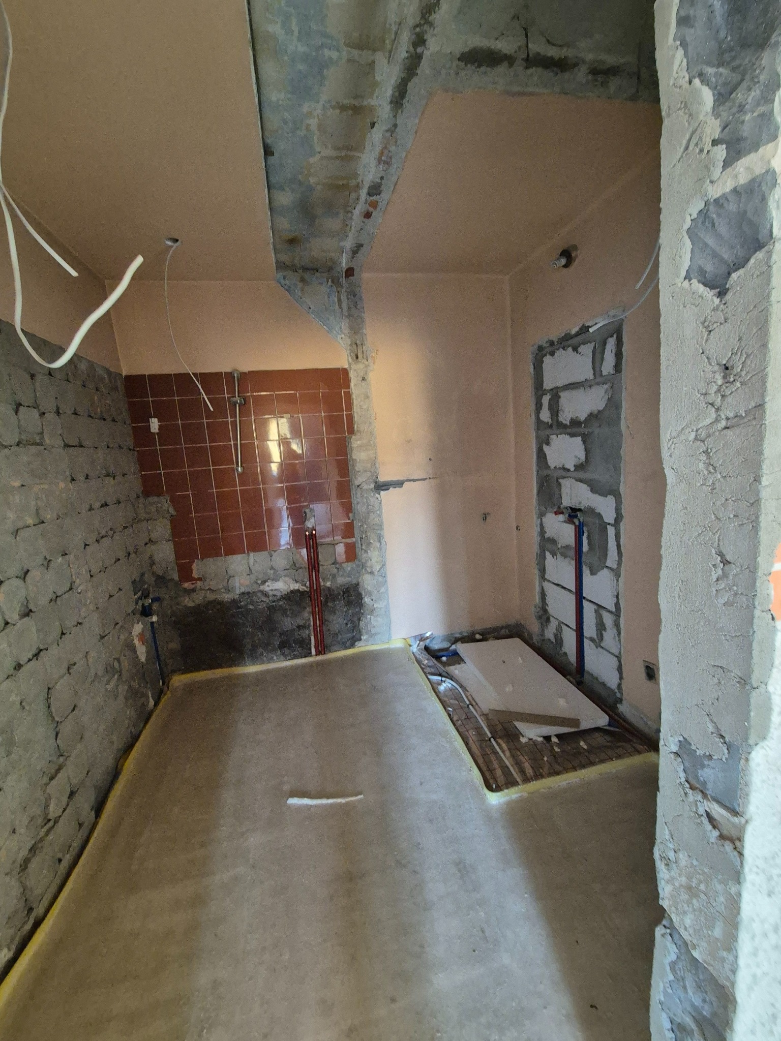 Wnętrze łazienki w trakcie prac remontowych: ściany częściowo obłożone brązowymi płytkami, fragmenty surowego muru, wystające rury, przygotowana instalacja ogrzewania podłogowego, widoczne przewody...