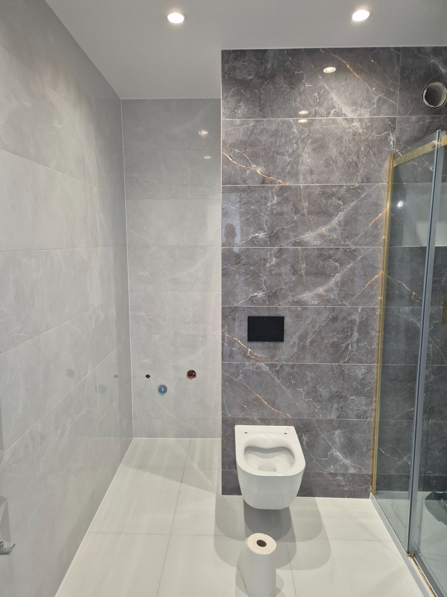 Nowoczesna łazienka z szarymi płytkami imitującymi marmur, podwieszana toaleta z czarnym przyciskiem spłukiwania, rolka papieru toaletowego na podłodze, kabina prysznicowa z elementami w kolorze...