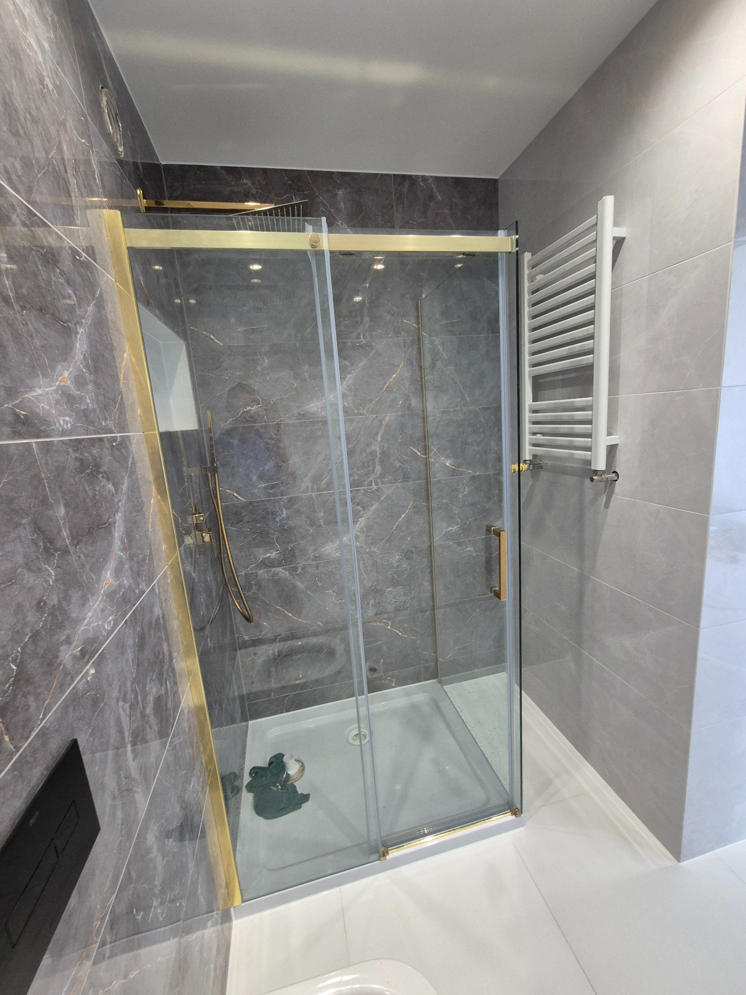 Nowoczesna kabina prysznicowa z przesuwnymi drzwiami, złotymi ramami i deszczownicą, na tle szarych płytek imitujących marmur, z białym grzejnikiem drabinkowym po prawej stronie.