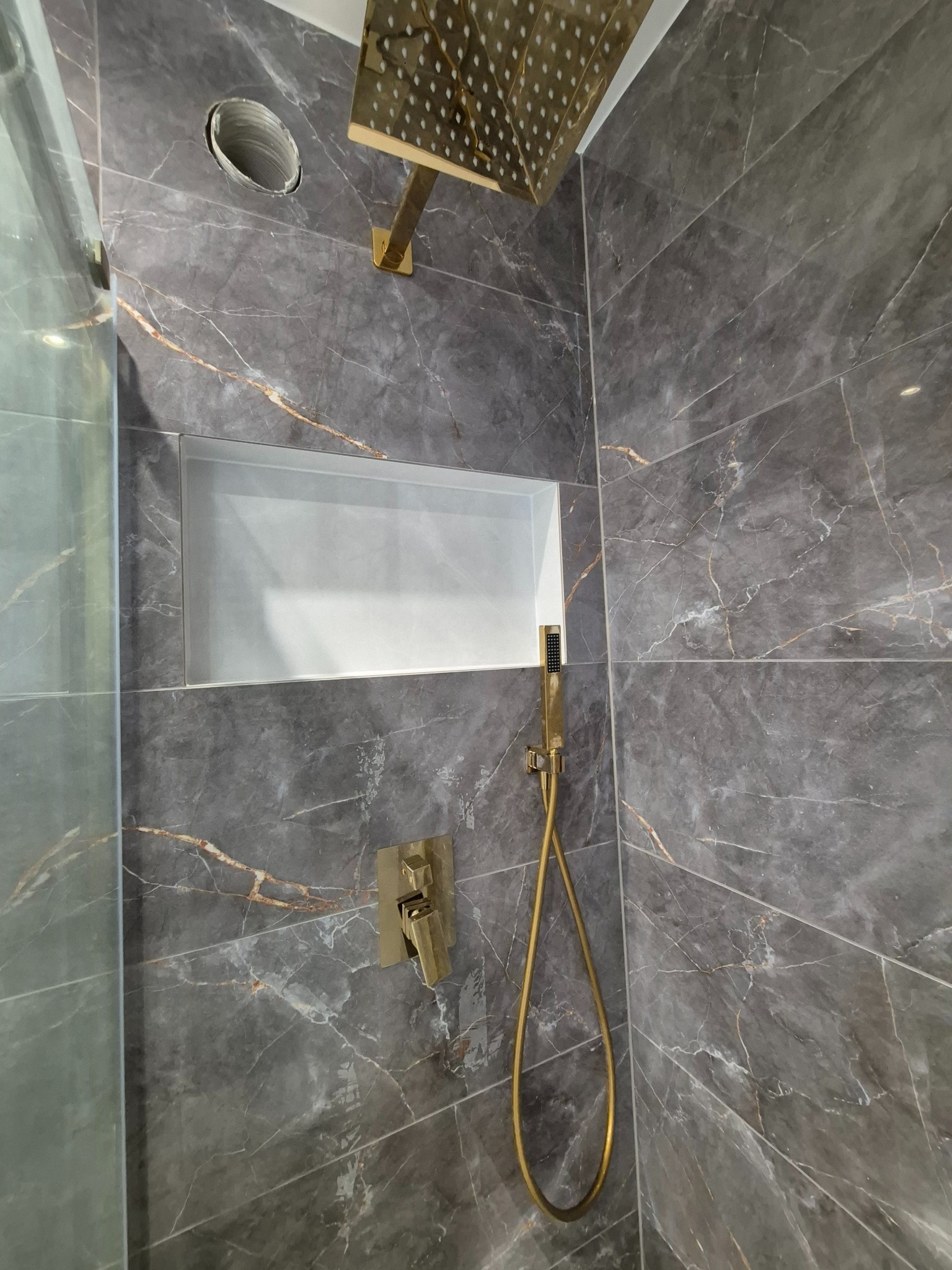 Szara łazienka wykończona płytkami imitującymi marmur z złotymi akcentami: deszczownica, bateria i słuchawka prysznicowa w kolorze złota. W ścianie wnęka z półką.