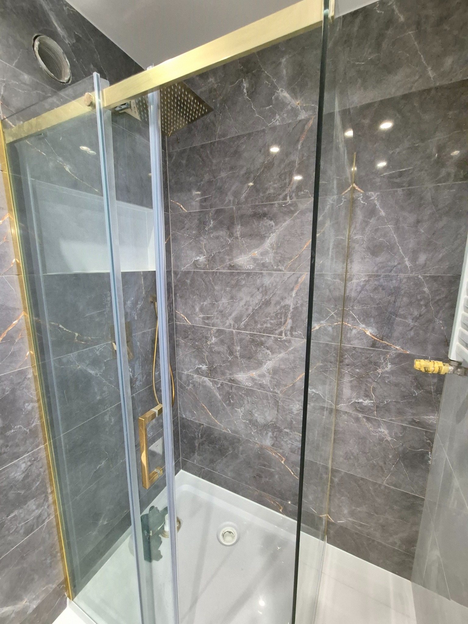 Szklana kabina prysznicowa z brodzikiem, złotymi ramami i deszczownicą, na tle marmurowych płytek w odcieniach szarości z delikatnym złotym żyłowaniem. Widoczny odpływ liniowy i fragment grzejnika.