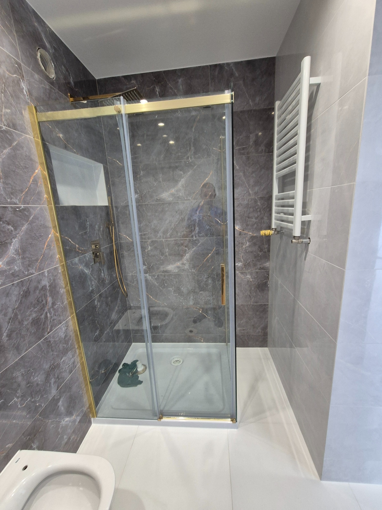 Nowoczesna kabina prysznicowa z złotymi elementami wykończeniowymi, marmurowe płytki na ścianach, biały grzejnik drabinkowy i fragment toalety w wyremontowanej łazience.