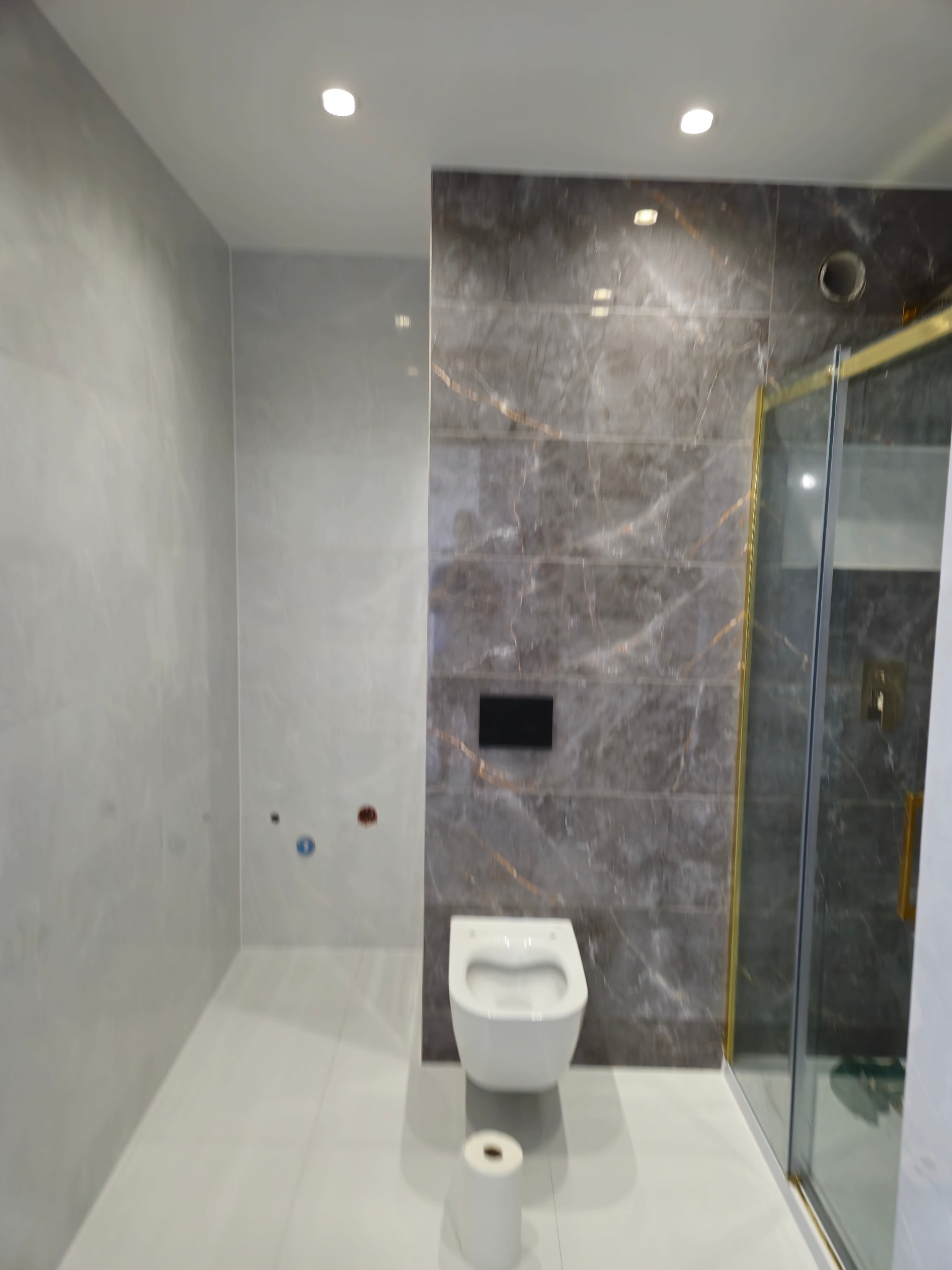 Nowoczesna łazienka po remoncie z marmurowymi płytkami, kabiną prysznicową ze złotymi ramami i podwieszaną toaletą. Widoczny również podajnik na papier toaletowy.