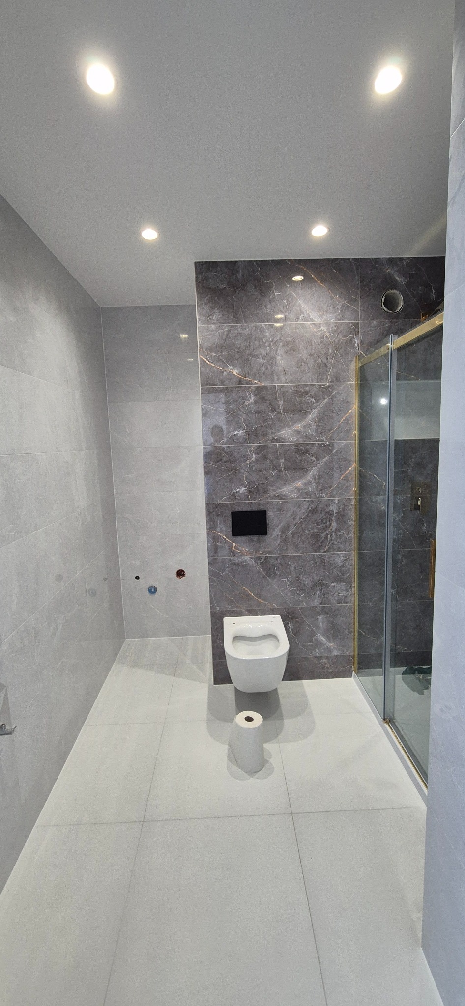 Nowoczesna łazienka z szarymi płytkami imitującymi marmur, podwieszana toaleta i kabina prysznicowa ze złotymi profilami; widoczny papier toaletowy na podłodze.