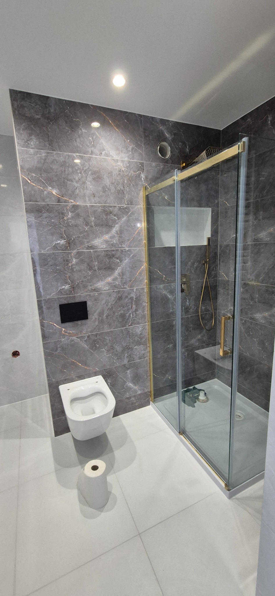 Nowoczesna łazienka z szarymi płytkami imitującymi marmur, kabiną prysznicową ze złotymi profilami i podwieszaną toaletą z czarnym panelem sterującym, uzupełniona o rolkę papieru toaletowego...