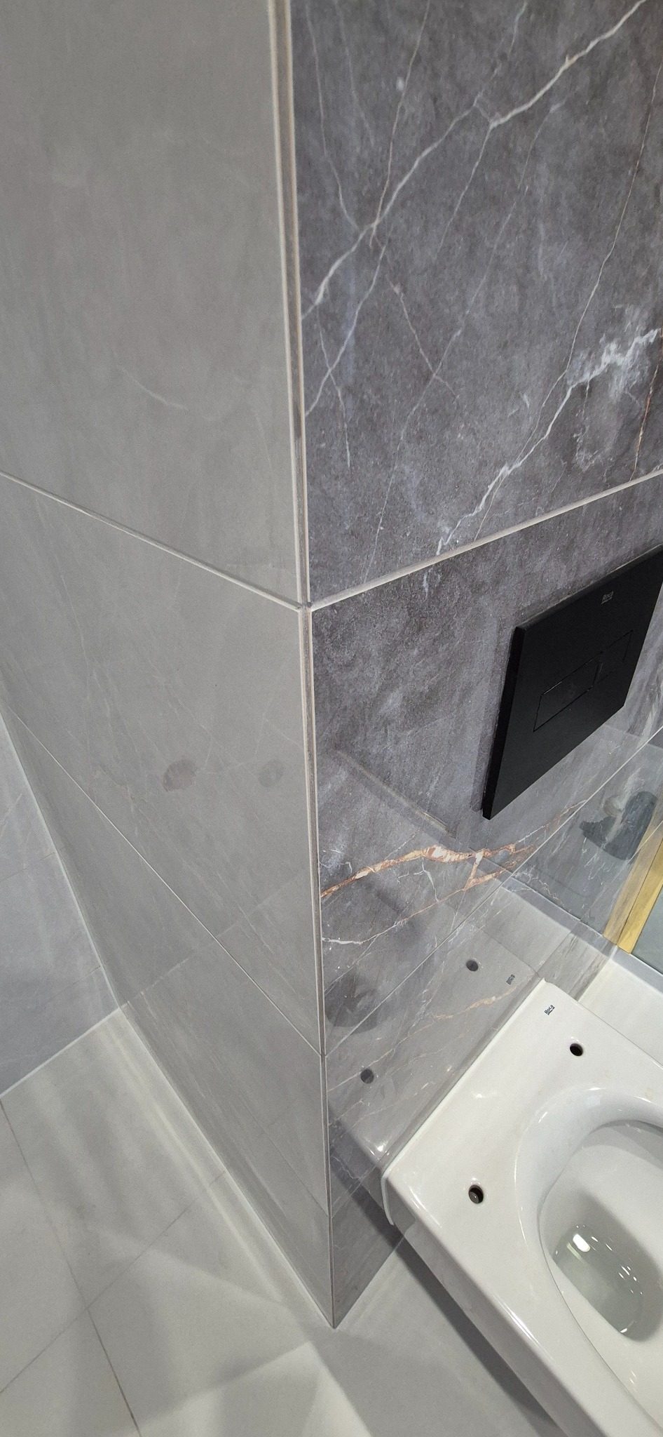 Fragment łazienki z szarymi płytkami imitującymi marmur, widoczny narożnik, panel sterujący spłuczką i biała miska WC w trakcie montażu, z widocznymi otworami na śruby.