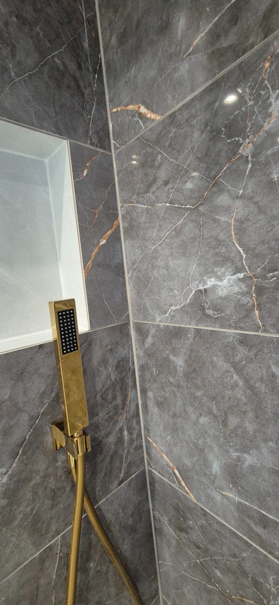 Błyszcząca, złota słuchawka prysznicowa na tle szarych, marmurkowych płytek w kabinie natryskowej z wnęką.