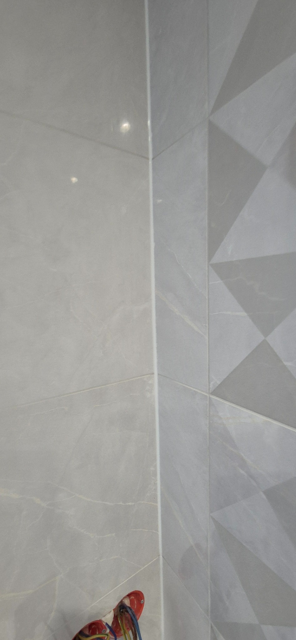 Szare płytki ścienne w łazience, gładkie i z geometrycznym wzorem, z widocznymi przewodami w dolnym rogu.