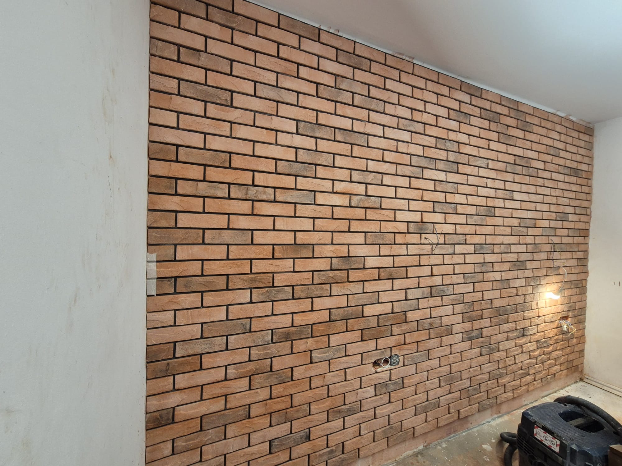 Ściana z cegieł dekoracyjnych w odcieniach brązu i beżu, z widocznymi puszkami elektrycznymi i prowizorycznym oświetleniem, prawdopodobnie w trakcie prac wykończeniowych w Myszkowie. W rogu...