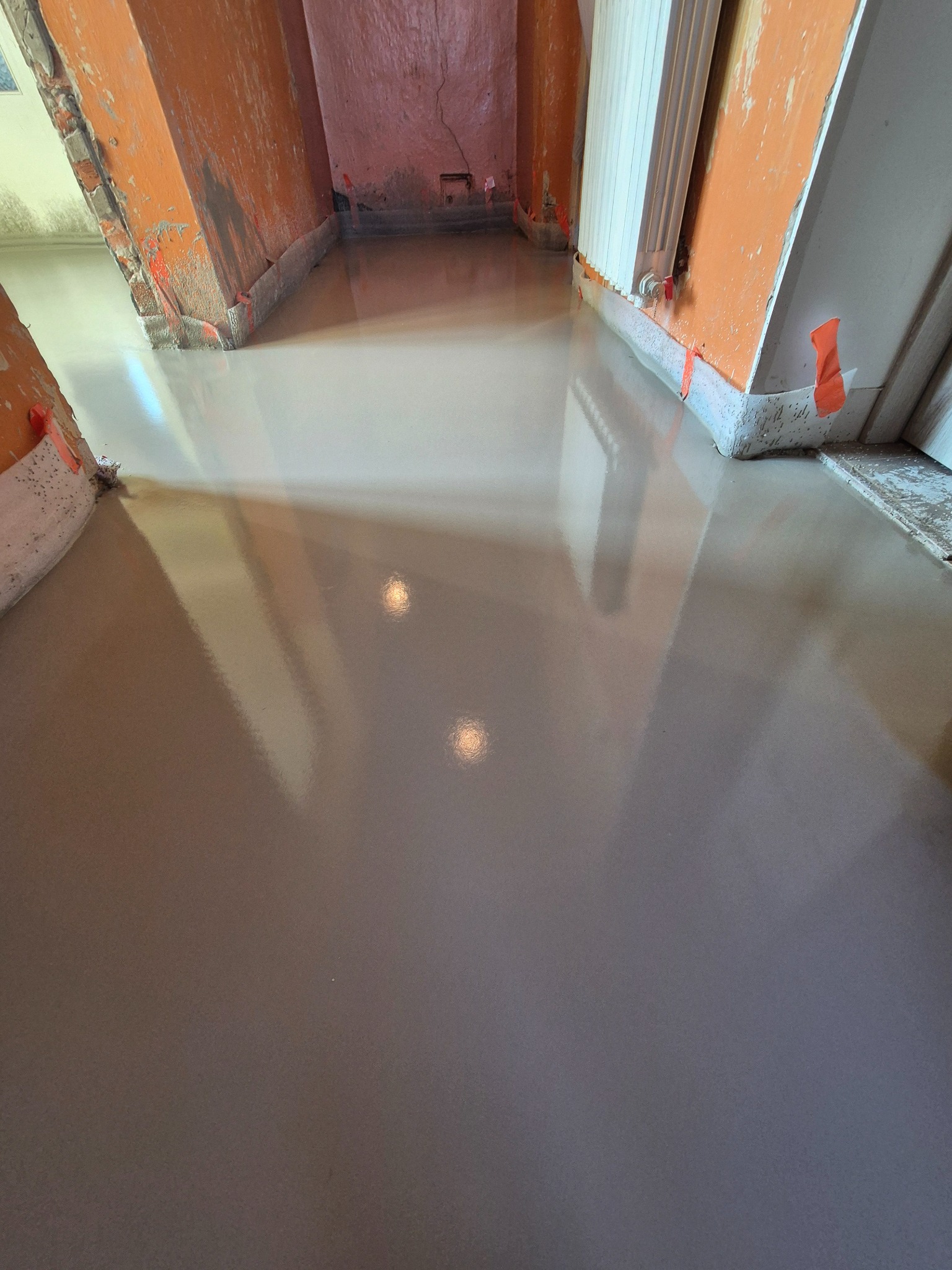 Świeżo wylana, samopoziomująca wylewka podłogowa w trakcie schnięcia, z widocznymi śladami taśmy malarskiej na ścianach i futrynach oraz odbiciami światła w mokrej powierzchni.