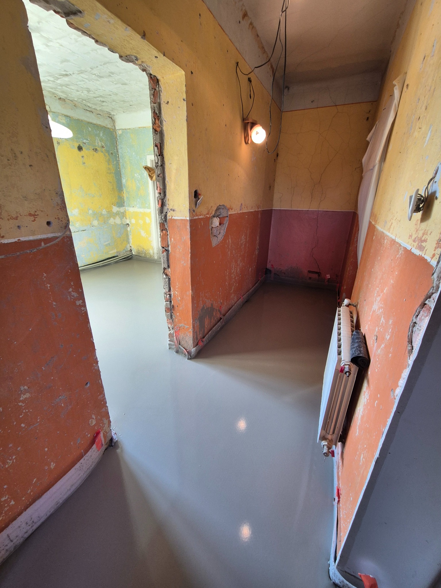 Wnętrze w trakcie wyrównywania podłogi wylewką samopoziomującą, widoczne stare, zniszczone ściany z odpadającą farbą w odcieniach żółci i pomarańczy oraz grzejnik.
