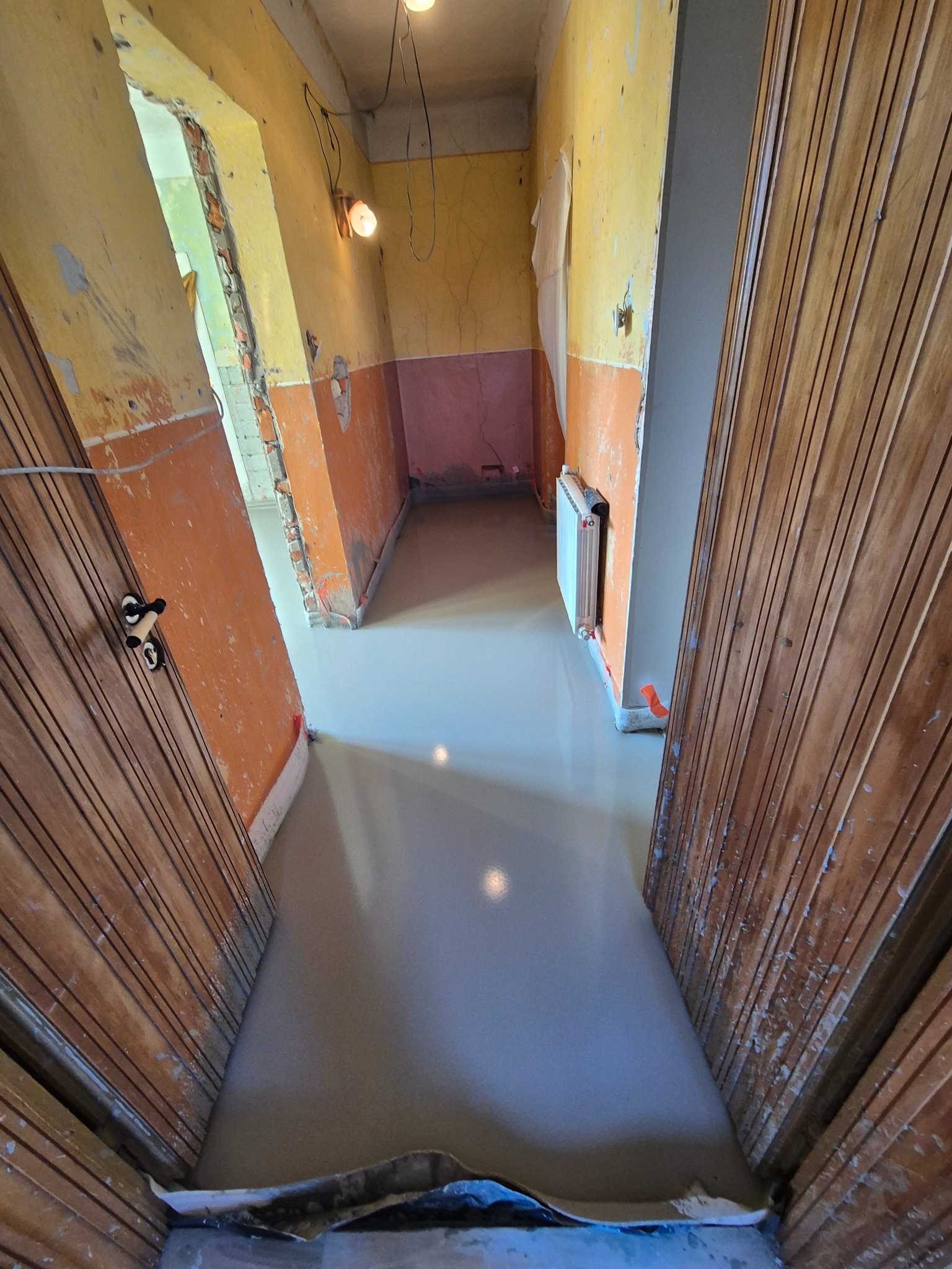 Wnętrze w trakcie prac wykończeniowych: wylewka na podłodze, ściany z fragmentami starej farby i tynku, widoczne drzwi z boazerią i grzejnik