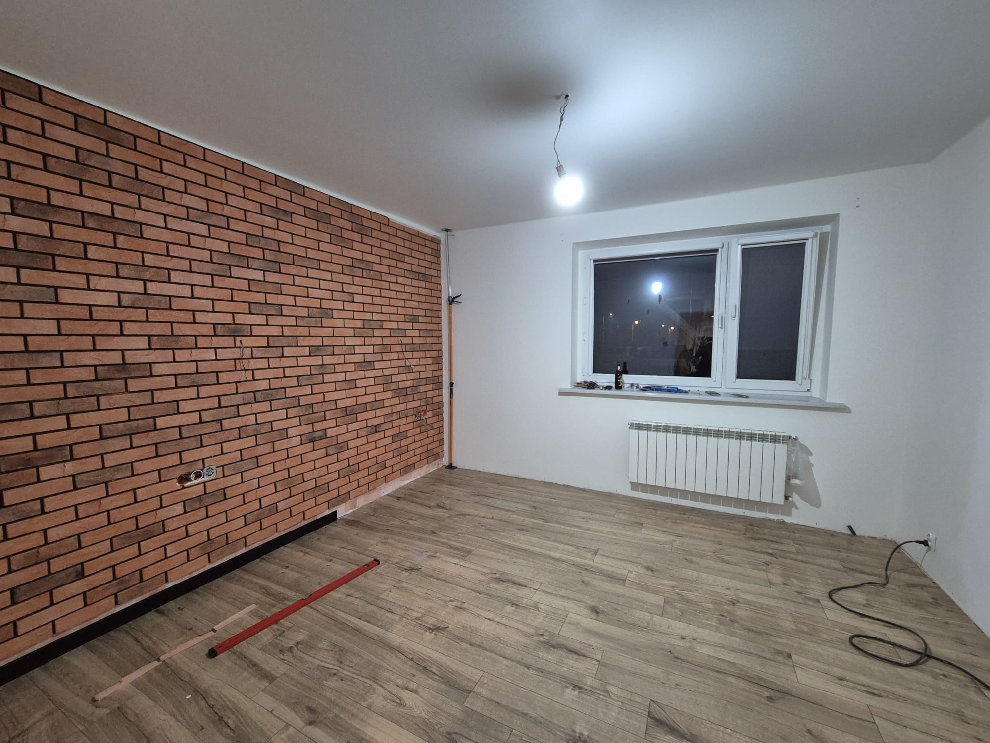 Pokój w trakcie prac wykończeniowych: ściana z cegły klinkierowej, panele podłogowe w odcieniach brązu, grzejnik pod oknem, wisząca żarówka i poziomica na podłodze.