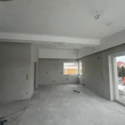 Surowe wnętrze w trakcie prac przygotowawczych, ściany pokryte szarą gładzią, widoczne punkty elektryczne, przewody wystające z sufitu, okna z widokiem na zimowy krajobraz.