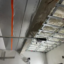 Sufit w trakcie prac: metalowa konstrukcja podwieszana, izolacja pianką natryskową, folia aluminiowa z pomarańczową taśmą uszczelniającą, widoczne przewody elektryczne.