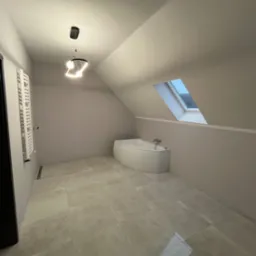 Nowoczesna łazienka na poddaszu z asymetryczną wanną, grzejnikiem drabinkowym i oknem dachowym, wykończona płytkami w odcieniach beżu, oświetlona designerską lampą.