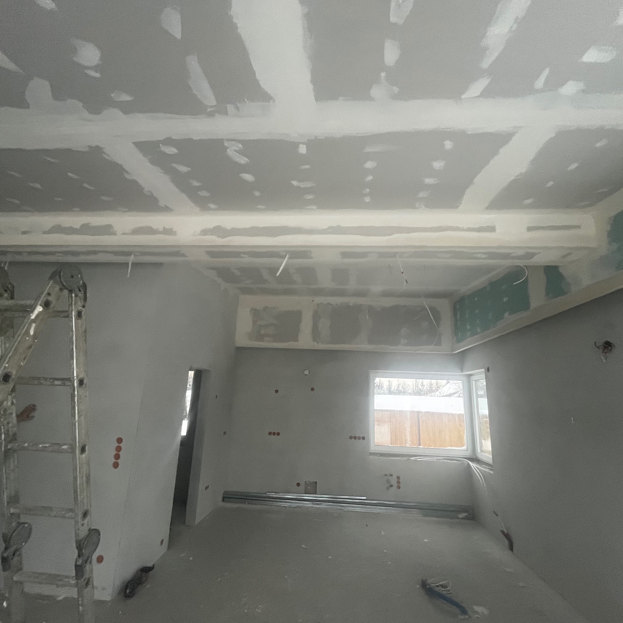 Wnętrze w trakcie prac: sufit z płyt kartonowo-gipsowych, częściowo zaszpachlowany, widoczna drabina i profile pod płytami, ściany przygotowane do dalszych prac wykończeniowych.