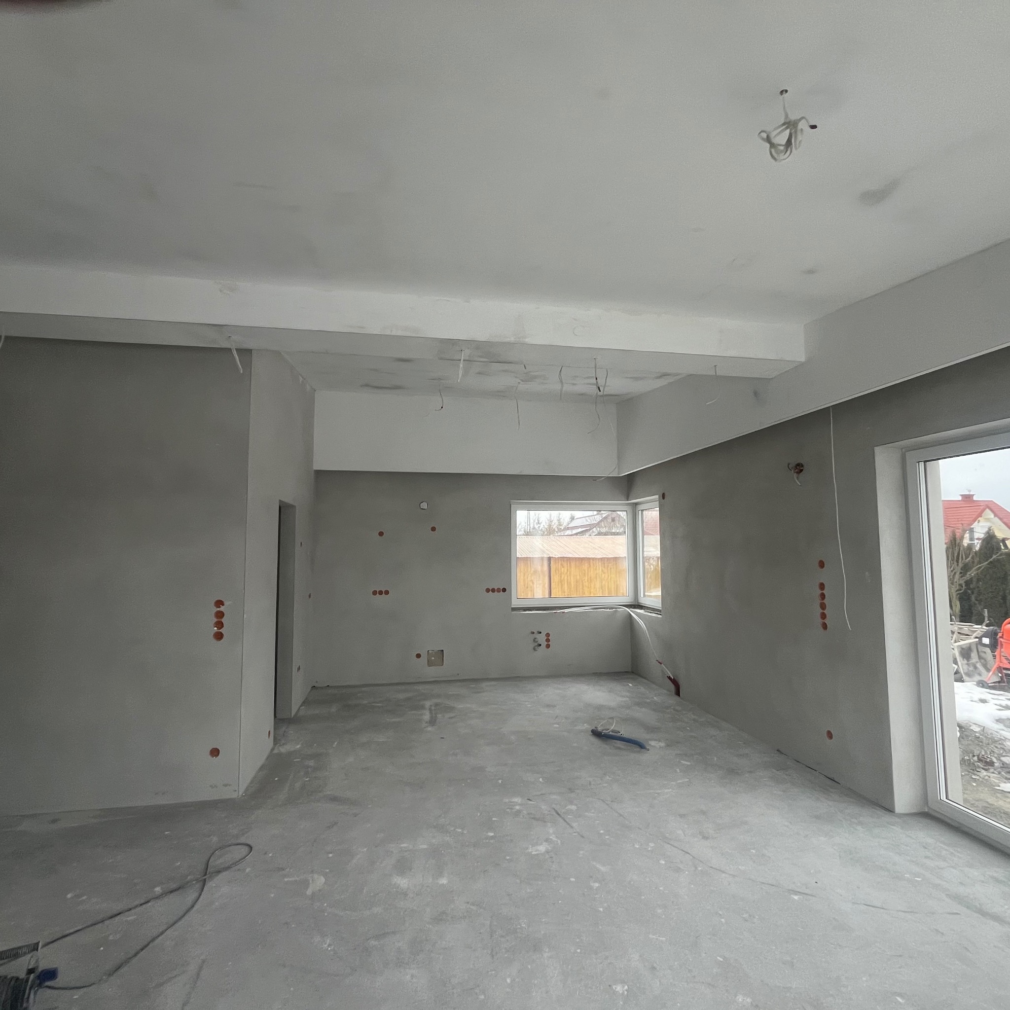 Surowe wnętrze w trakcie prac przygotowawczych, ściany pokryte szarą gładzią, widoczne punkty elektryczne, przewody wystające z sufitu, okna z widokiem na zimowy krajobraz.