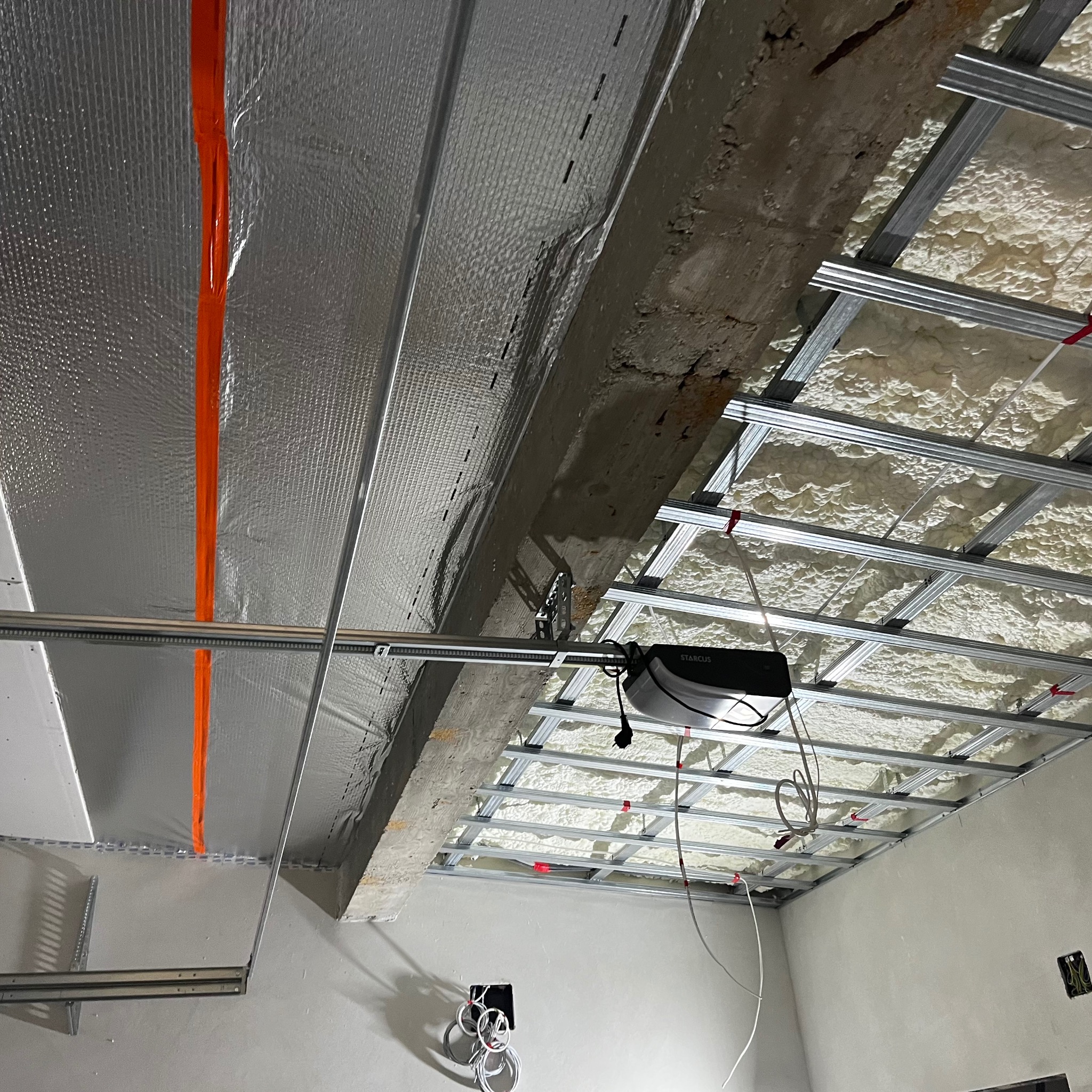 Sufit w trakcie prac: metalowa konstrukcja podwieszana, izolacja pianką natryskową, folia aluminiowa z pomarańczową taśmą uszczelniającą, widoczne przewody elektryczne.