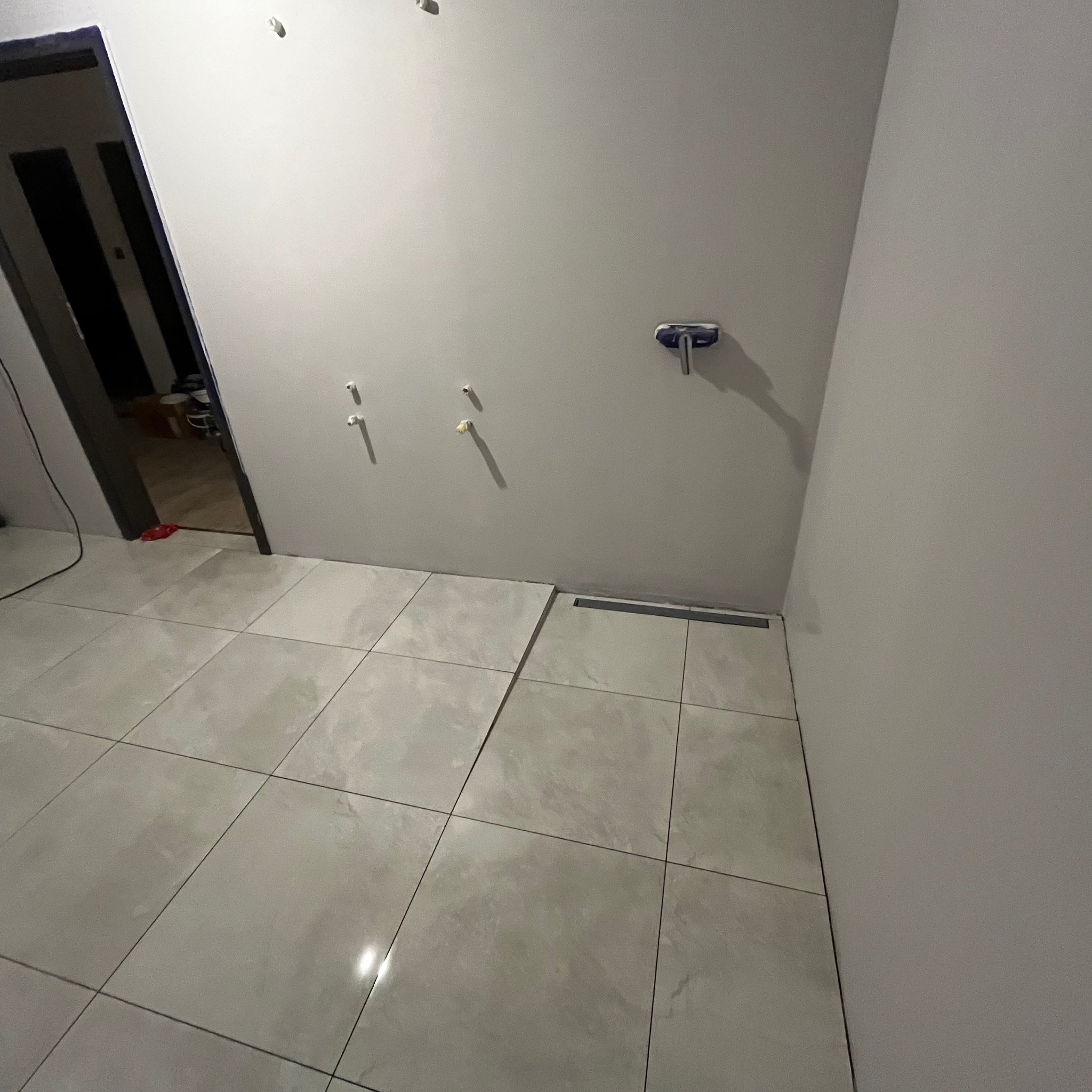 Wnętrze łazienki w trakcie remontu, z ułożonymi szarymi płytkami podłogowymi, widoczne wyprowadzenia instalacji wodnej na ścianie i odpływ liniowy.