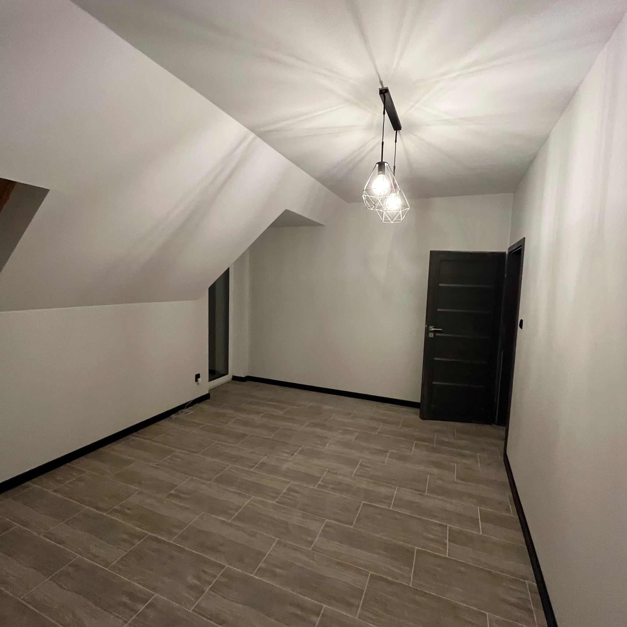 Poddasze po remoncie z drewnopodobną podłogą, białymi ścianami i czarnymi listwami przypodłogowymi oraz czarnymi drzwiami, oświetlone nowoczesną lampą wiszącą.