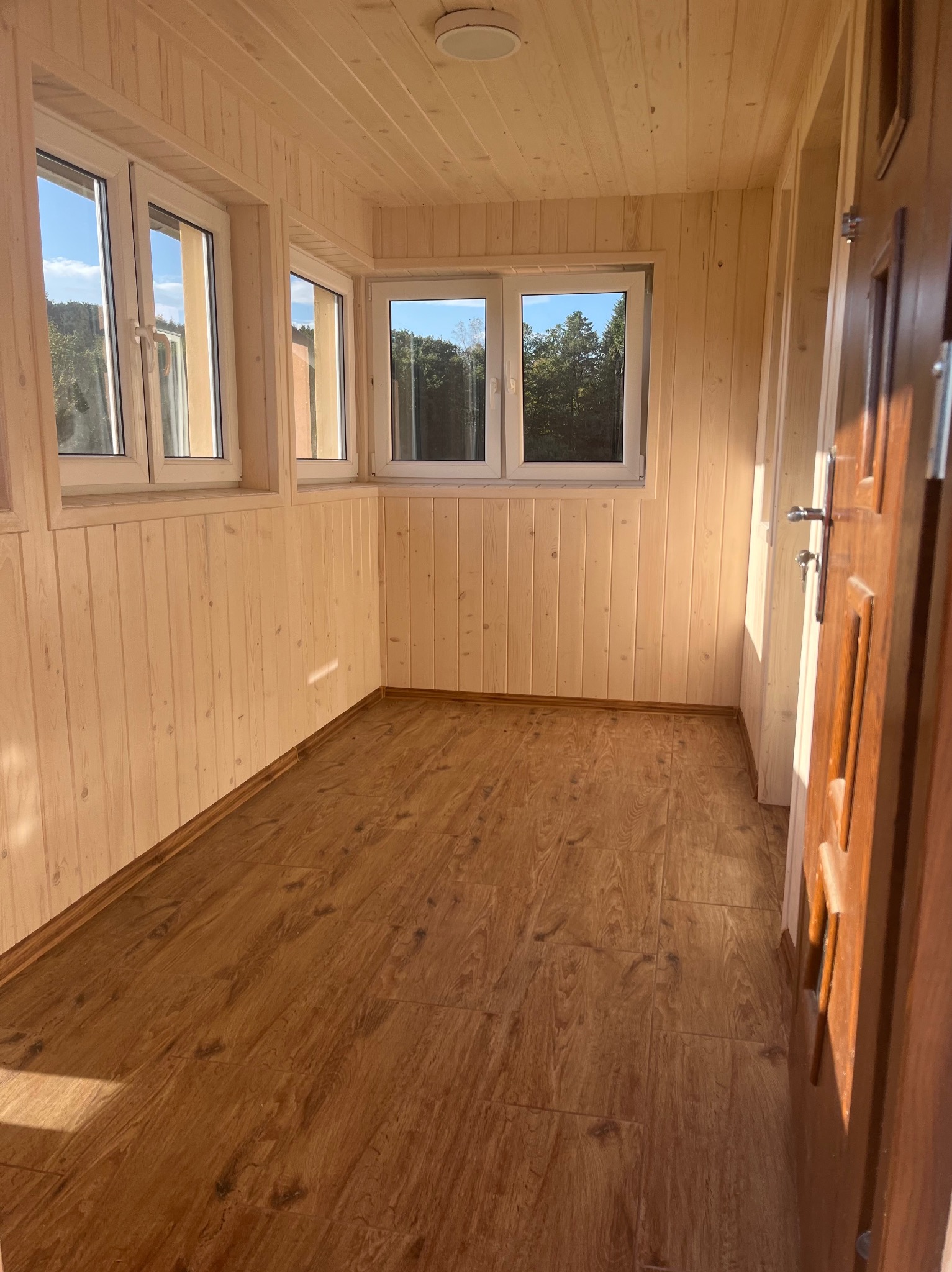 Jasne wnętrze wykończone boazerią i panelami podłogowymi w odcieniach brązu, z widocznymi trzema oknami i częściowo otwartymi drewnianymi drzwiami po prawej stronie, wpadające promienie słoneczne.