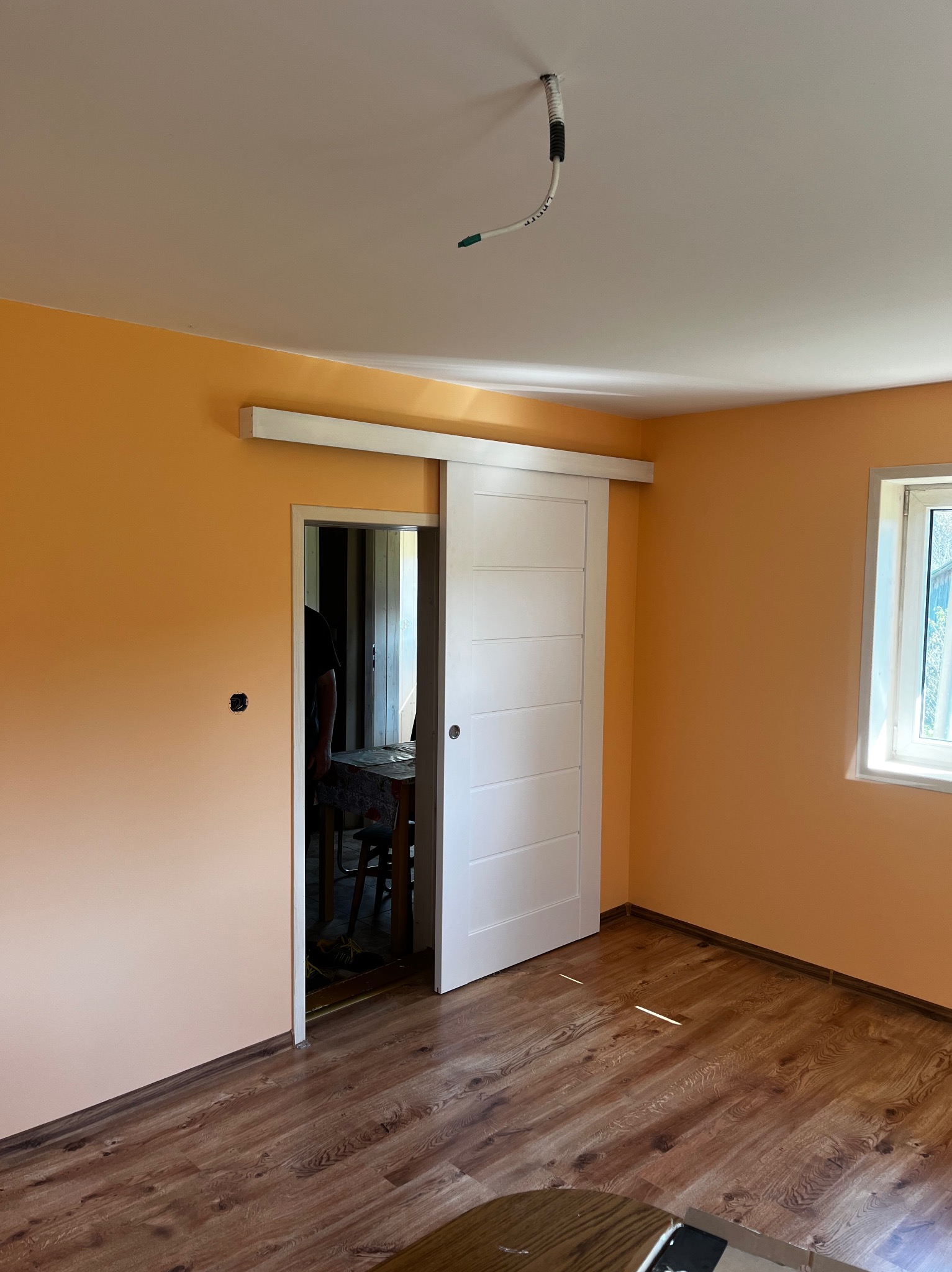 Jasne wnętrze pokoju z pomarańczowymi ścianami, drewnianą podłogą i przesuwnymi białymi drzwiami. Widoczny fragment kabla wystającego z sufitu oraz okno z widokiem na zieleń.